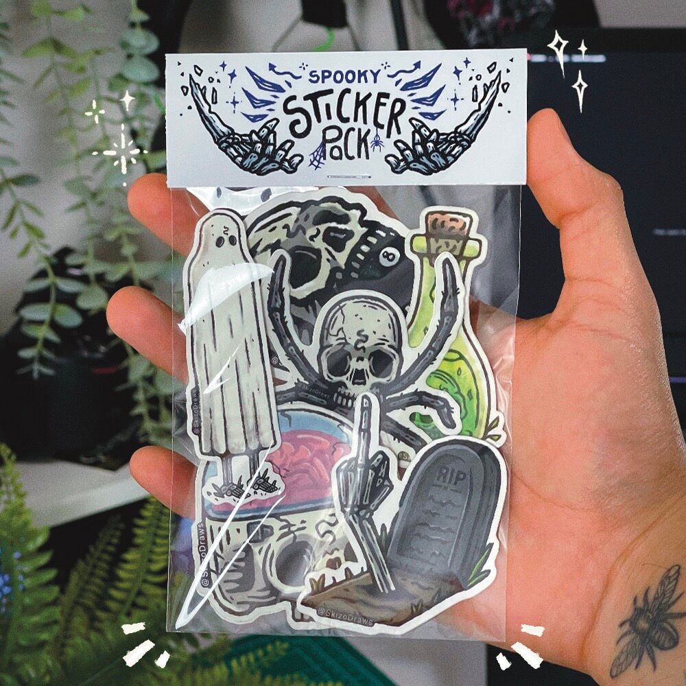Käsi pitelee tarrapakettia, jossa on kauhuteemaisia piirrostarroja ja pakkauskortti, jossa lukee "SPOOKY STICKER PACK". Taustalla kasveja.