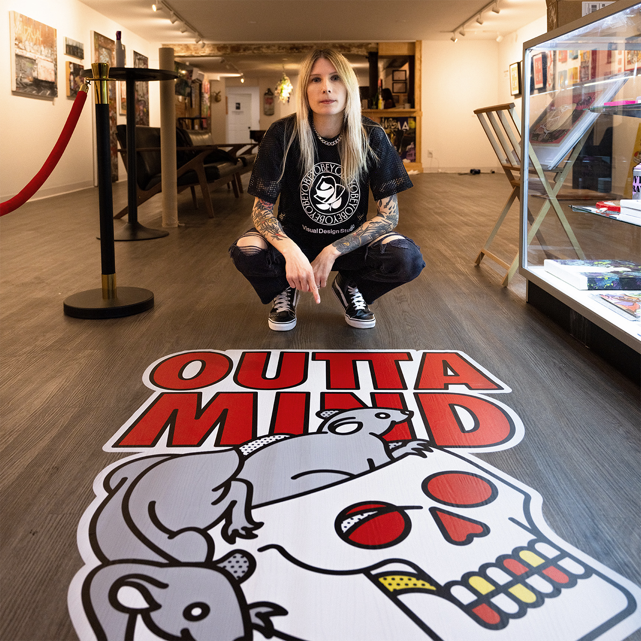 Henkilö polvistuu värikkään "OUTTA MIND"-tarran vieressä galleriassa, ympärillään taideteoksia.