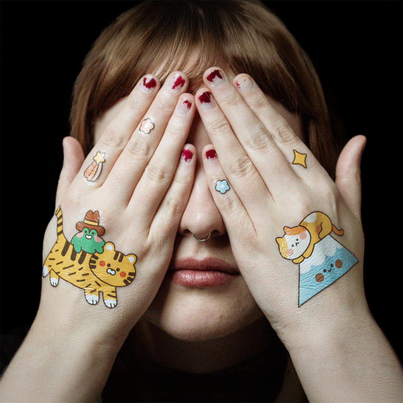 Person covering their face with both hands, decorated with colorful temporary tattoos of cute animals, stars, and small playful designs.