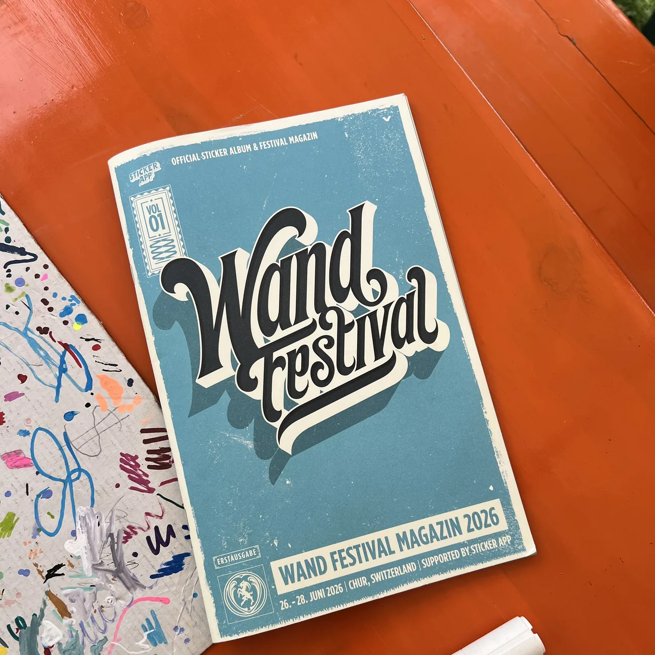 Wand Festivalin 2026 virallinen tarra-albumi ja festivaalilehti StickerAppin tukemana maalitussin vieressä oranssilla pöydällä