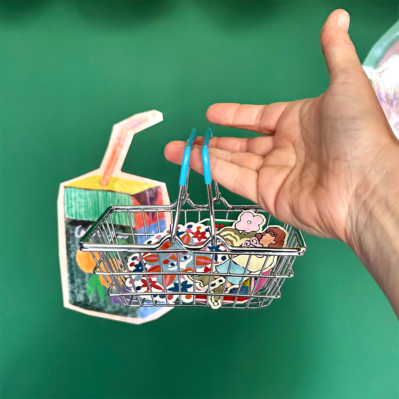 Hand holding a miniature shopping basket filled with colorful paper cutouts, next to a cartoon-style juice box on a green background.