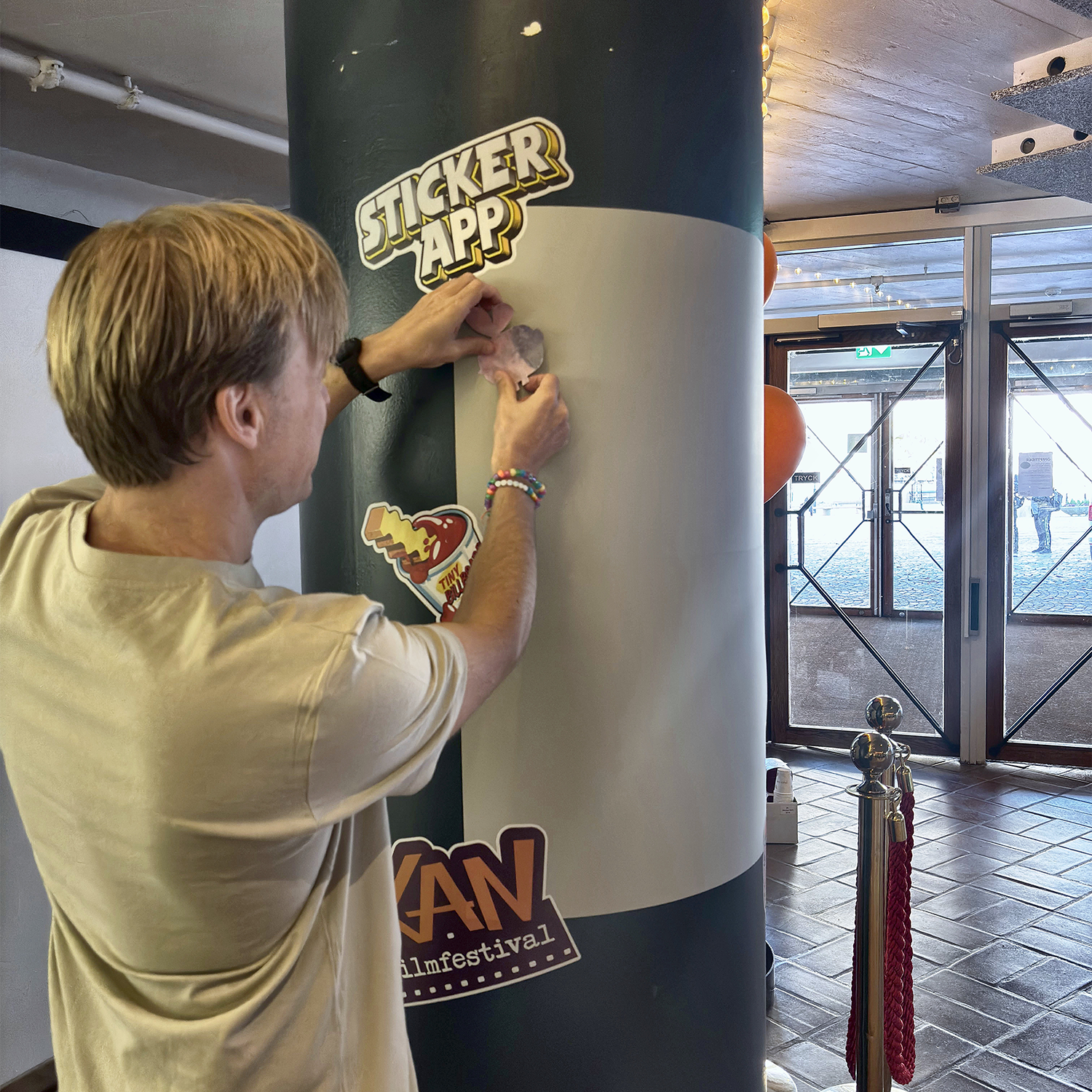 Mies kiinnittää tarraa pilarin ympärille kiedotulle StickerAppin tarraseinälle KAN Film Festival -tapahtumassa