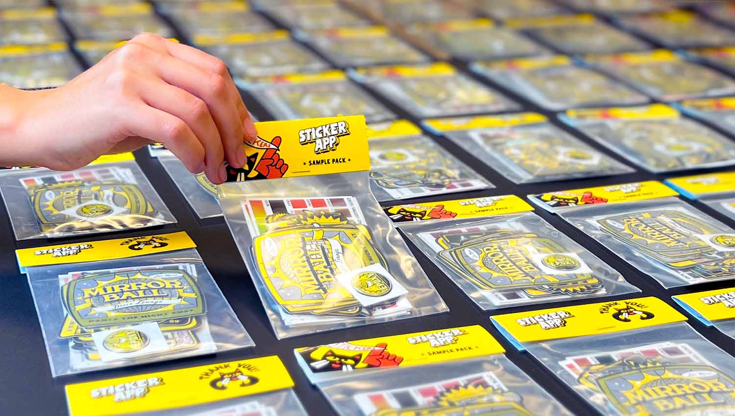 A hand lifts a sticker pack from a stack on a darkened table, highlighting the sample pack while others remain neatly arranged.