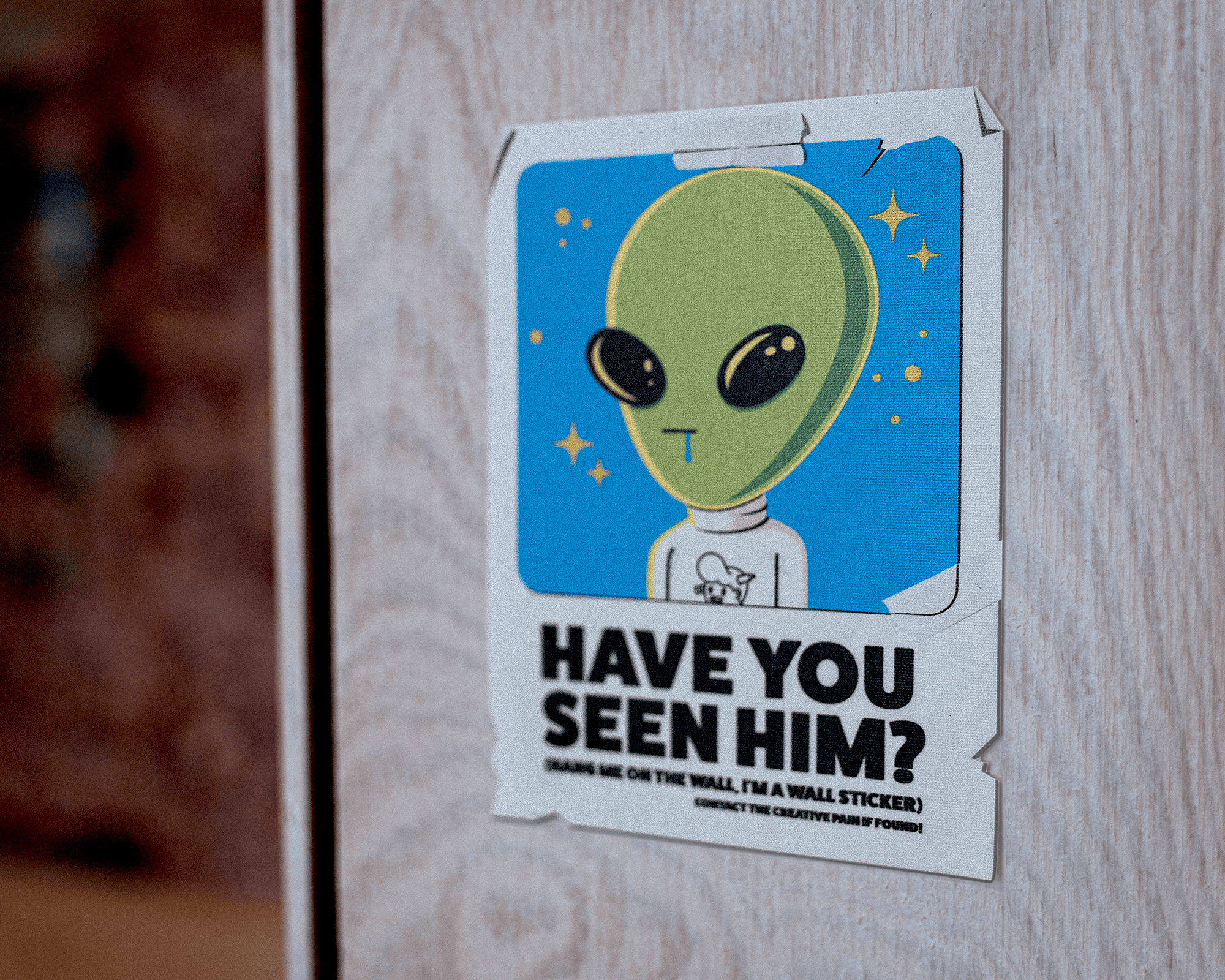 Wall sticker of a cartoon alien with large eyes on a blue background, captioned "Have you seen him?" on a wooden surface.