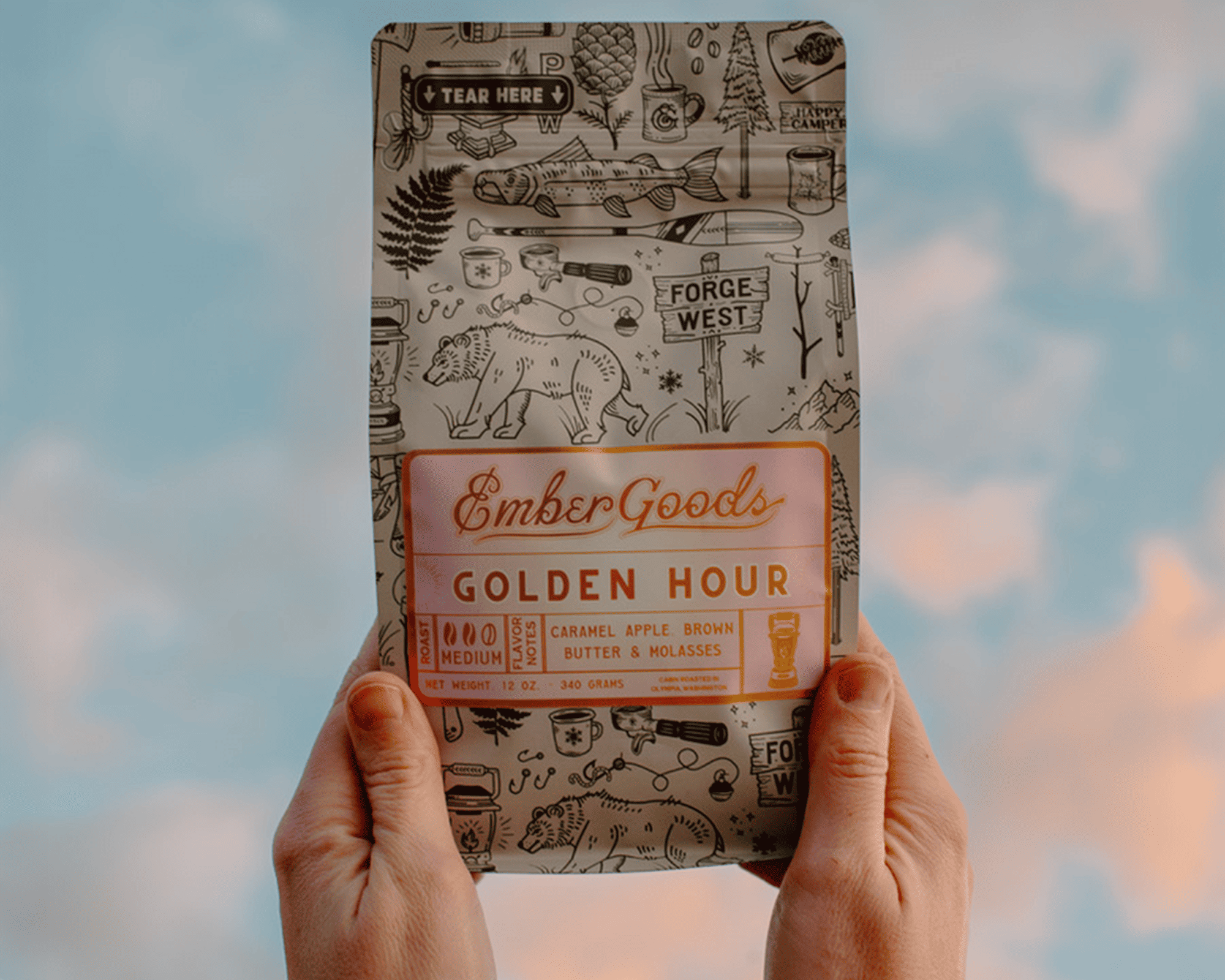 Kädet pitelevät Ember Goodsin Golden Hour -kahvipussia, jossa on metallinen etiketti, taustalla taivas