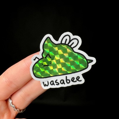 A hand holding a green Wasabee prismatic sticker with a face on it